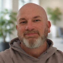 David smiles at the camera, he has a beard and is wearing a brown hoodie.