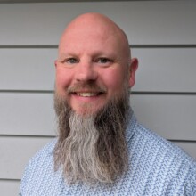 Jon smiles at the camera, he has a long beard and is wearing a light blue shirt