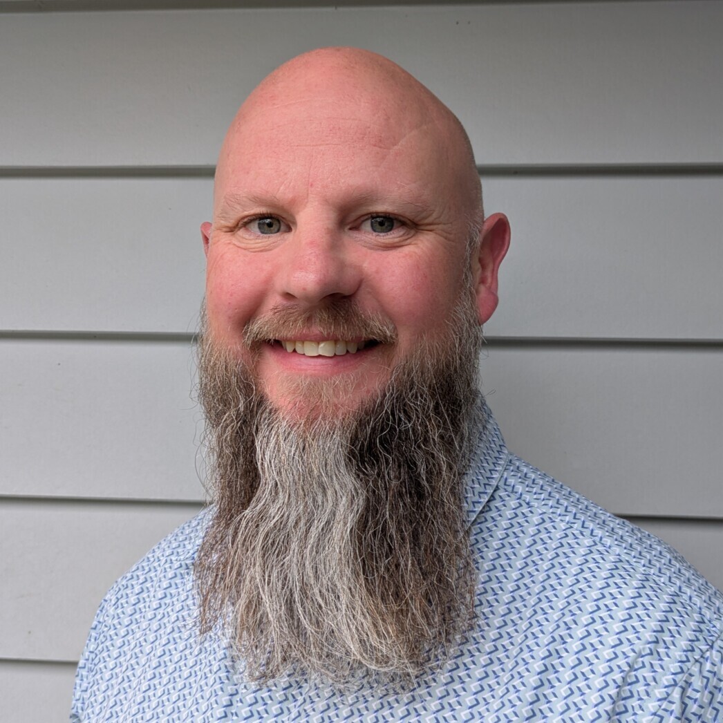 Jon smiles at the camera, he has a long beard and is wearing a light blue shirt