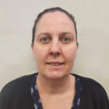 Al stands in front of a blank wall. Her dark hair is tied back and she is smiling.