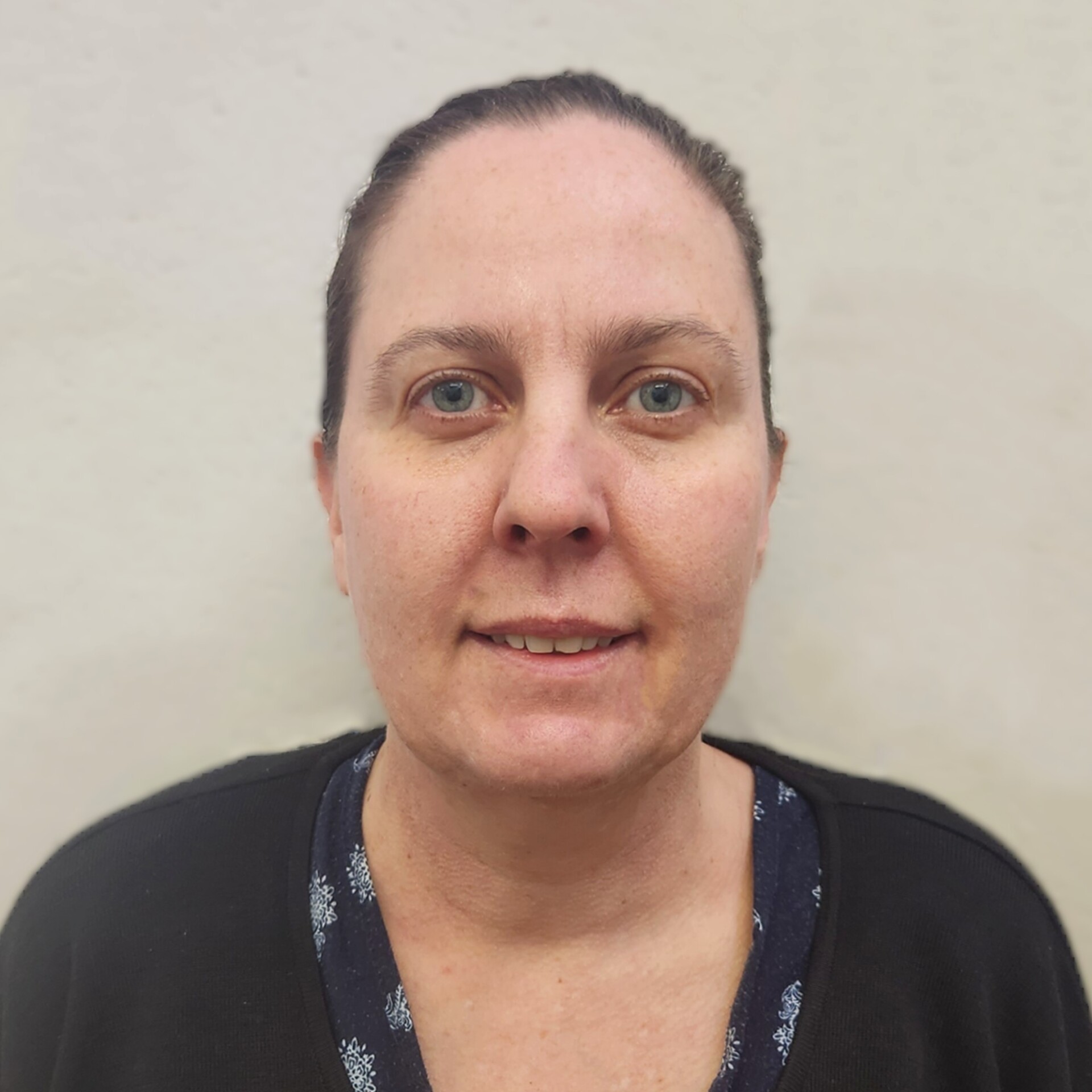 Al stands in front of a blank wall. Her dark hair is tied back and she is smiling.