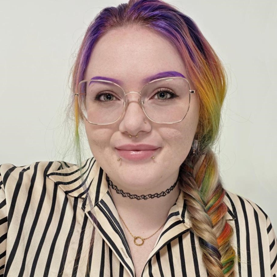 Morgan is smiling. She has rainbow coloured hair, purple eyebrows and is wearing reading glasses.