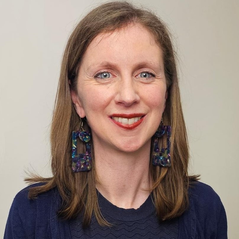 Alana's smile is framed by red lipstick, she has large colourful earrings and her shoulder-length hair is a brown-auburn colour.