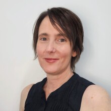 Eliisa smiles, her hair is short and dark, and she is wearing a dark top.