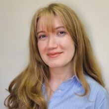 Esra smiles for the camera in front of a plain wall. Her full, blonde hair rests on her shoulders