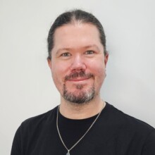 Gwyn smiles beneath a moustache and beard, his long dark hair is tied back, and he is wearing a black shirt and silver chain.