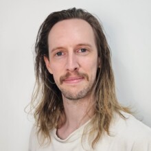 Simon smiles. He has long dark-to-light hair, and facial hair, he is wearing a light shirt.