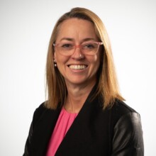 Jane has shoulder length light brown hair is wearing glasses and a pink top under a black jacket