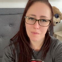 Claire has shoulder length brown hair is wearing glasses and a grey shirt with a pic on the front