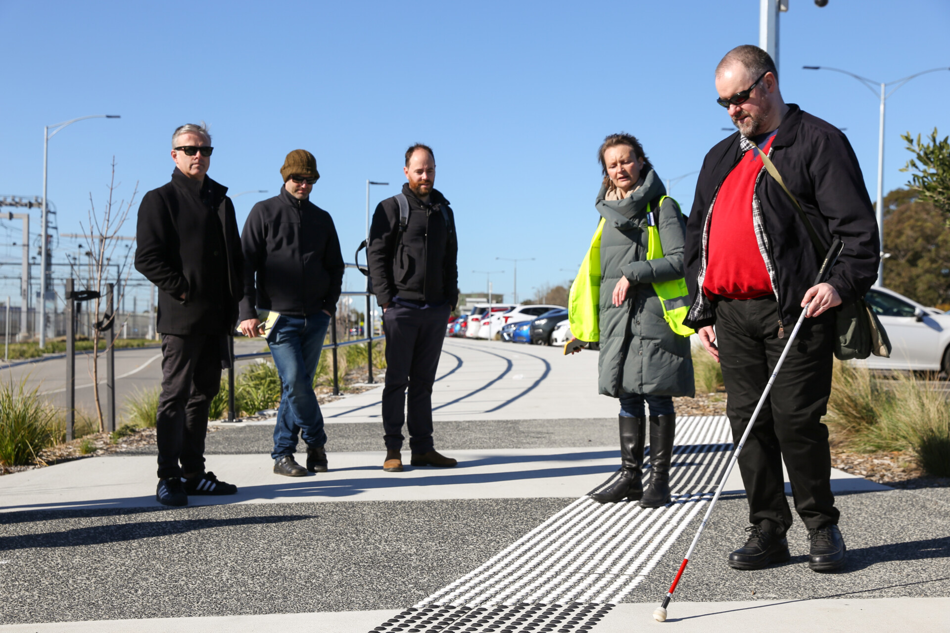 A group of people observe and discuss as a white cane user tests how their cane reacts to tactile strips on the footpath.
