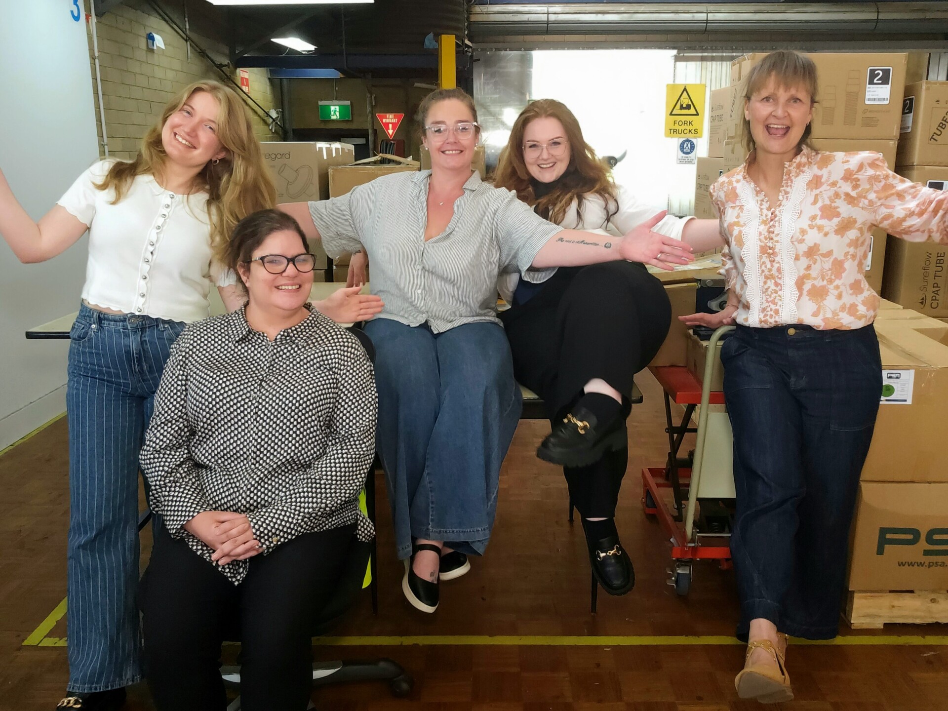 Five women pose for a photo in the Ability Works assembly area. They are smiling and have their arms extended outwards.