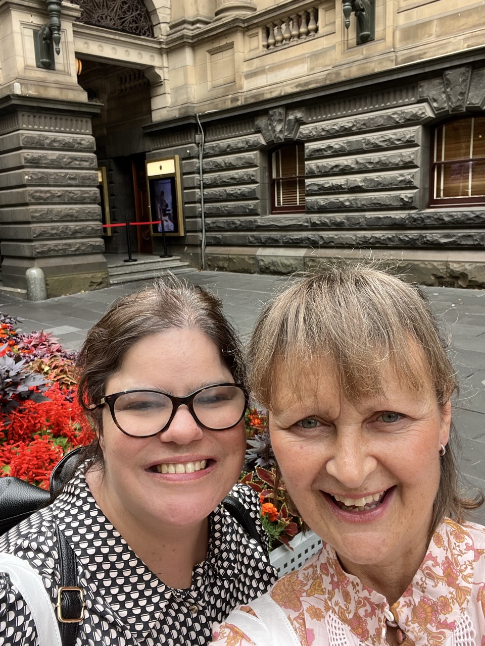 Ruby and Natalie smile for a selfie in front of a stone building and flower beds.