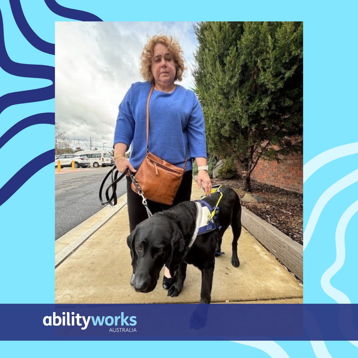 Debra has light, curly hair. She is standing on a foot path holding Royce's harness. Royce is her dark coloured seeing eye dog.