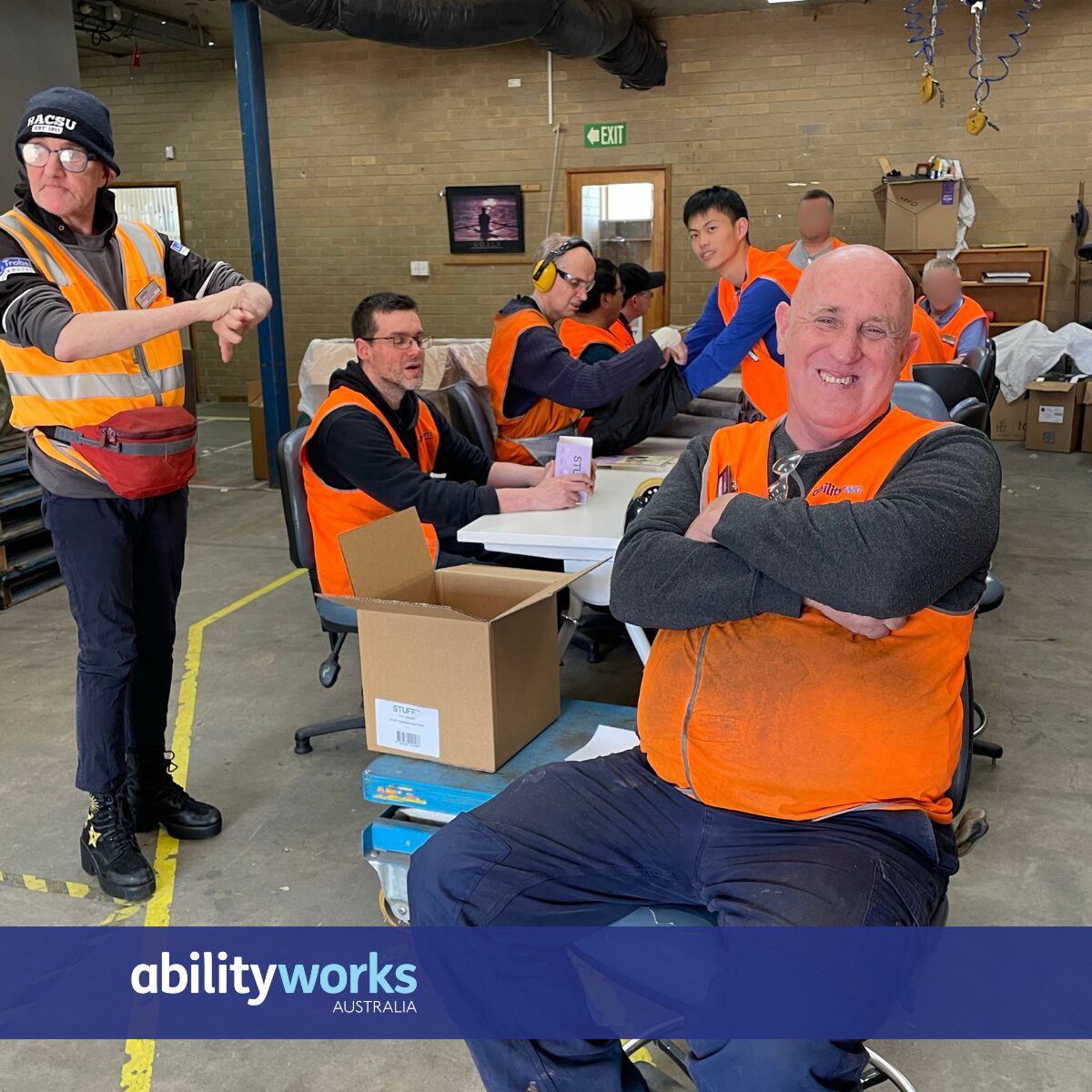 Several men in orange safety vests are gathered in a warehouse setting, with one in the foreground smiling with his arms crossed.